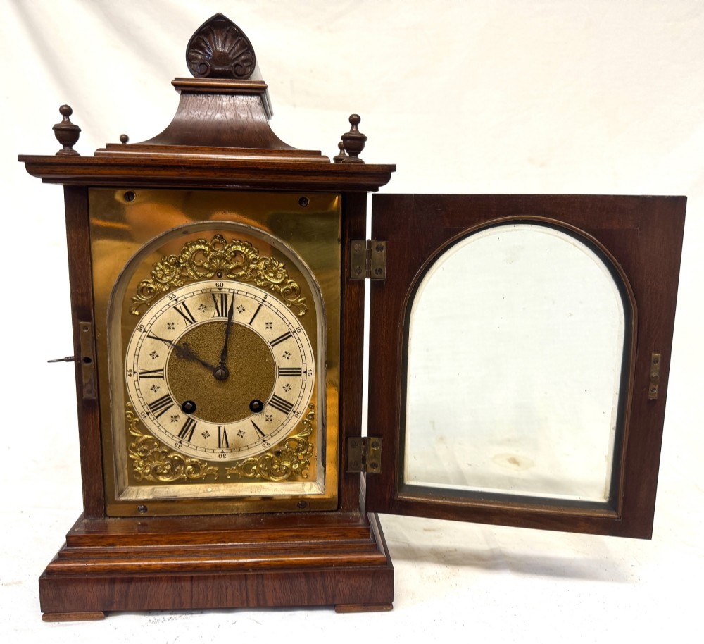 An antique walnut bracket clock featuring an arched brass dial with decorative corner spandrels, Roman numerals, and turned finials on a stepped wood base. View III