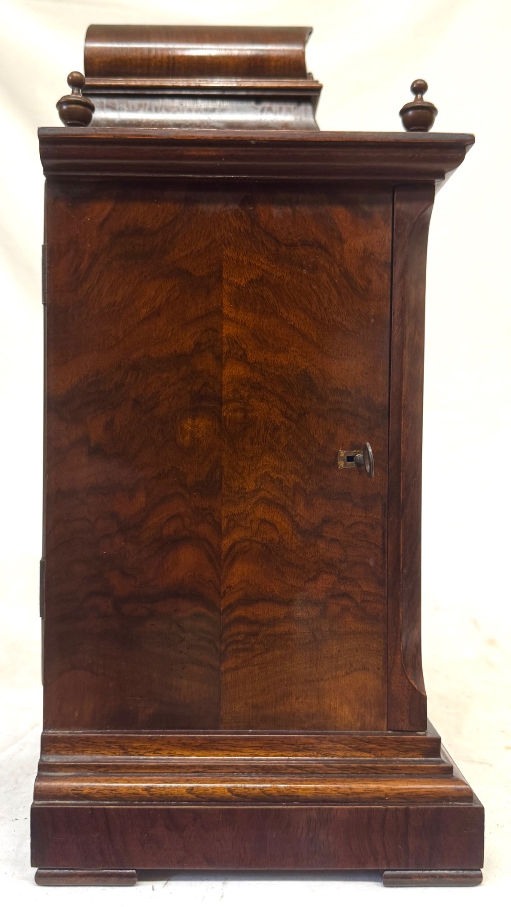 An antique walnut bracket clock featuring an arched brass dial with decorative corner spandrels, Roman numerals, and turned finials on a stepped wood base. View V