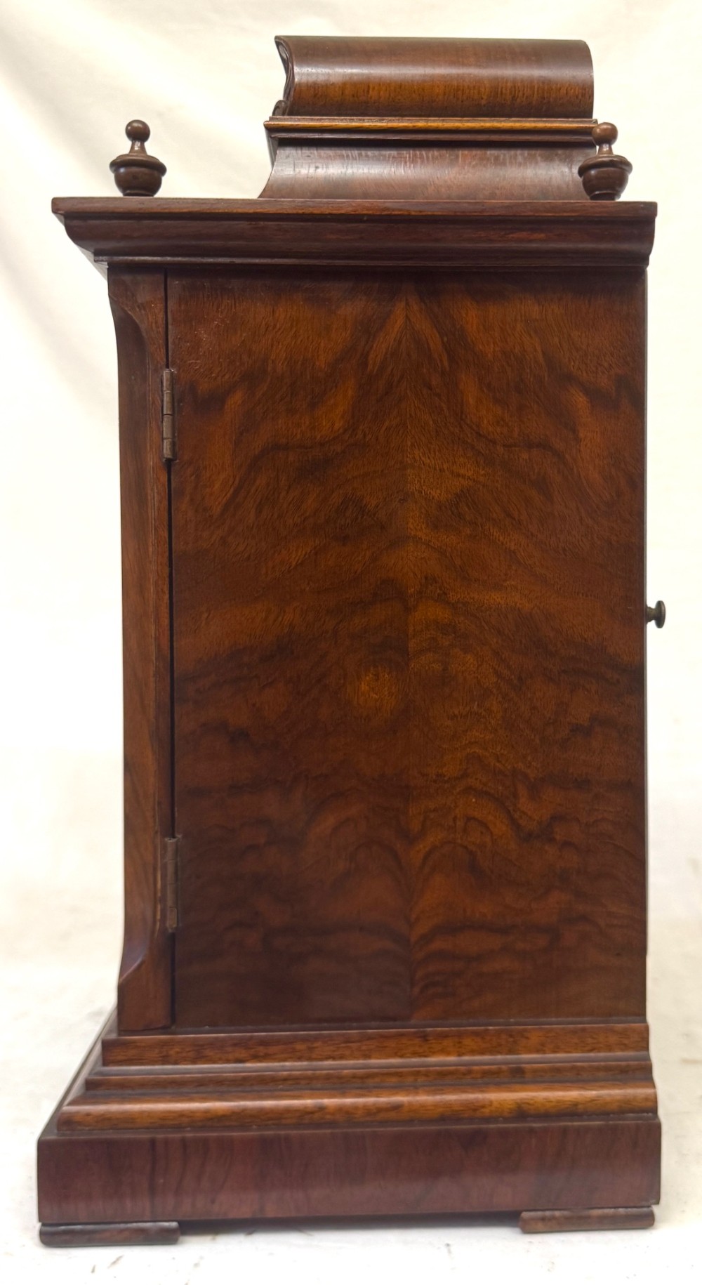 An antique walnut bracket clock featuring an arched brass dial with decorative corner spandrels, Roman numerals, and turned finials on a stepped wood base. View VI