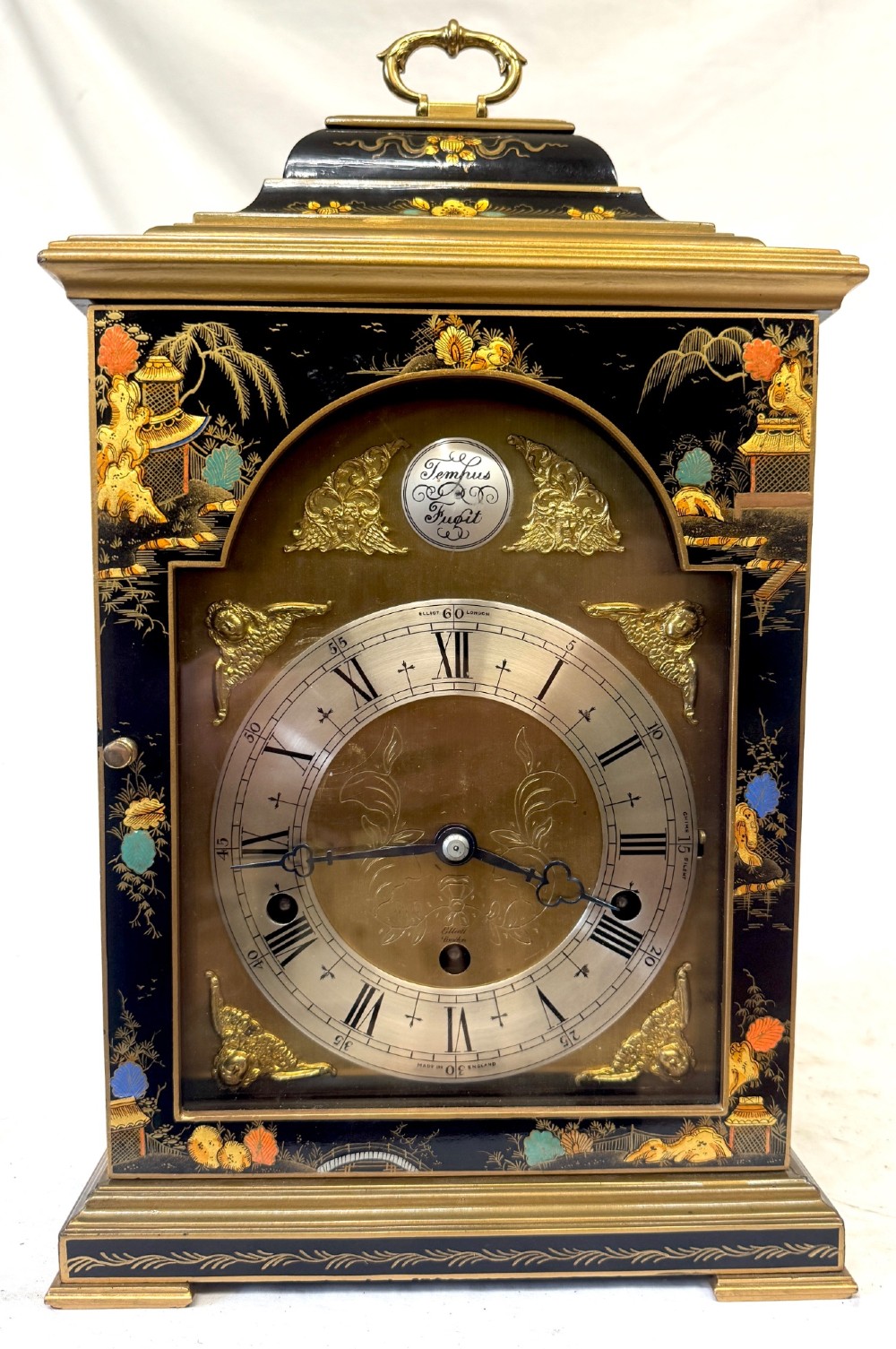 A black lacquered chinoiserie bracket clock featuring hand-painted gilt landscape scenes, a brass dial with Roman numerals, and a top brass carrying handle. View II