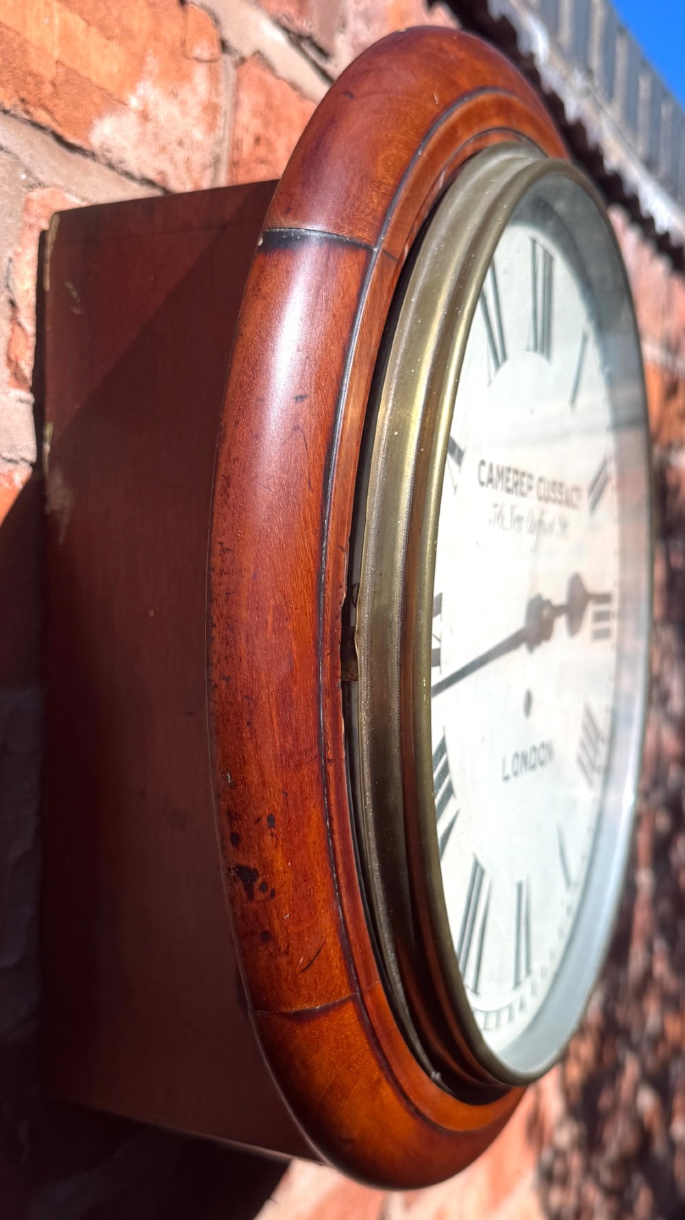 A circular Victorian mahogany wall clock with a cream painted dial displaying Roman numerals, makers inscription for Camerer Cuss & Co London, and a brass bezel. View VI