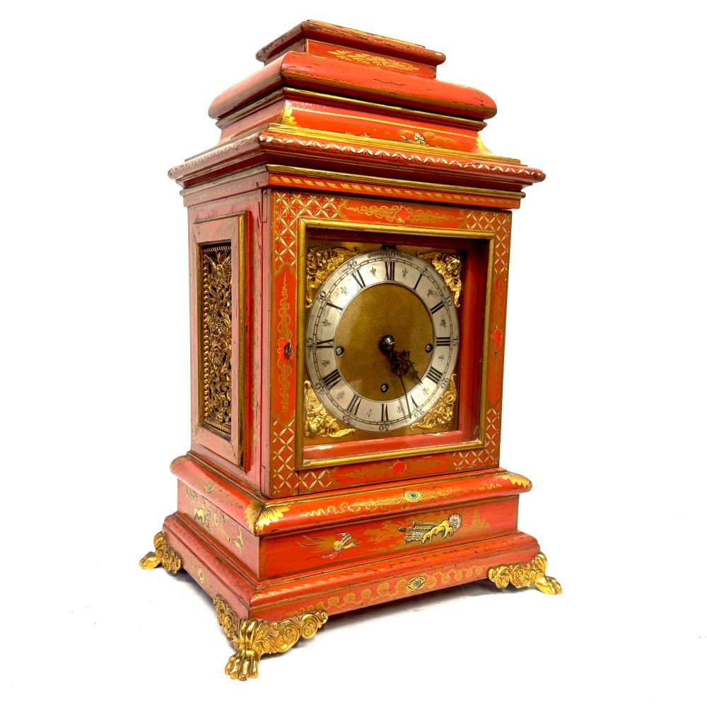 A red lacquered bracket clock decorated with gold chinoiserie motifs, featuring a brass dial and gilt metal feet.