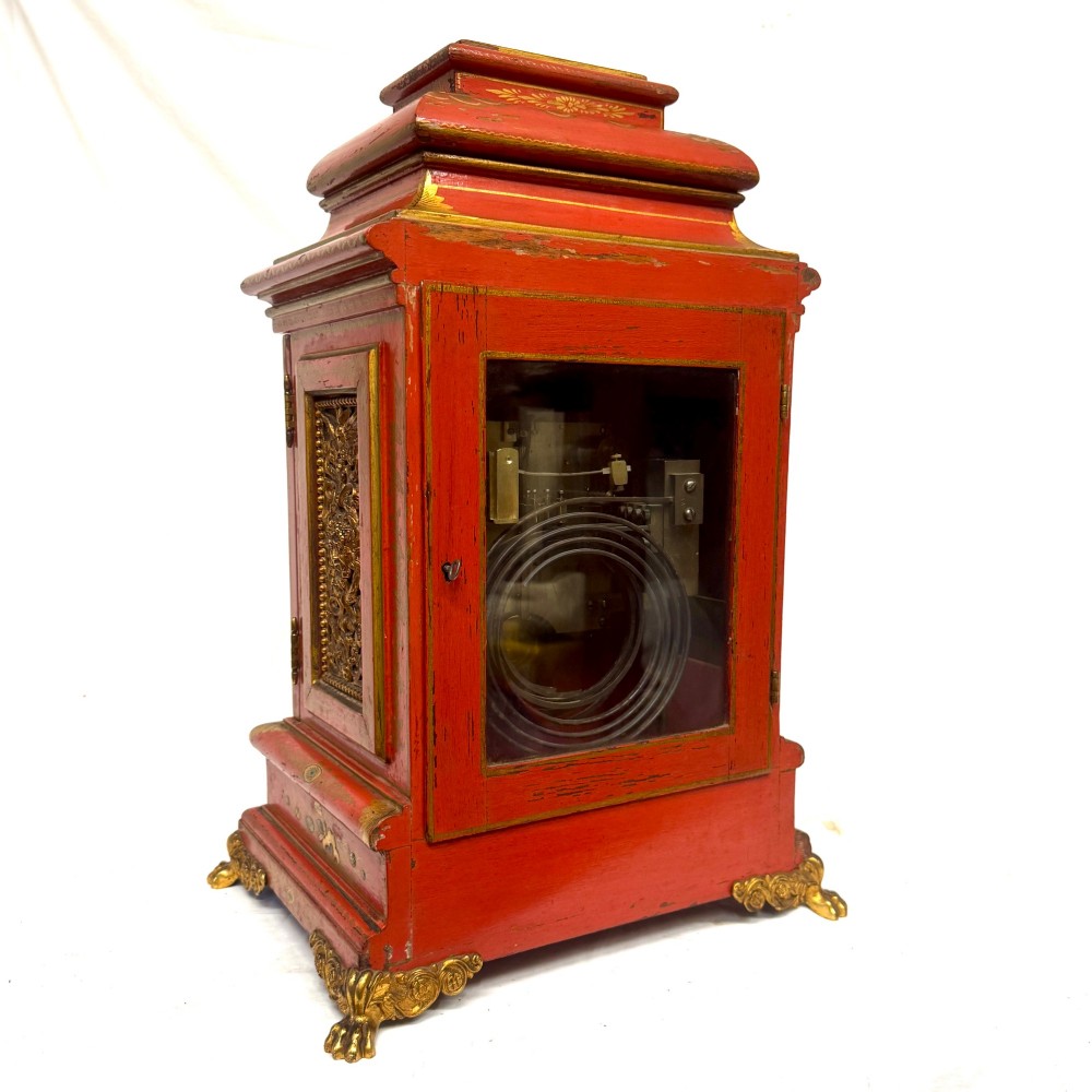 A red lacquered bracket clock decorated with gold chinoiserie motifs, featuring a brass dial and gilt metal feet. View V