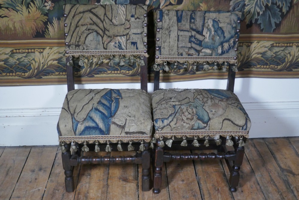 A pair of 17th-century oak back stools upholstered in tapestry fabric with tassel trim and turned legs.