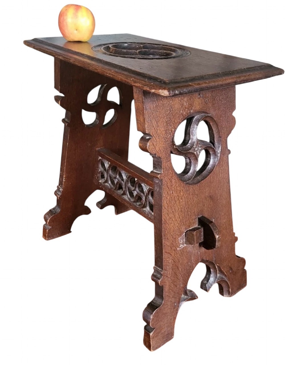 A 19th-century dark oak Gothic Revival stool featuring carved tracery cut-outs on the slab legs, a carved central stretcher, and a carved recessed motif on the top.