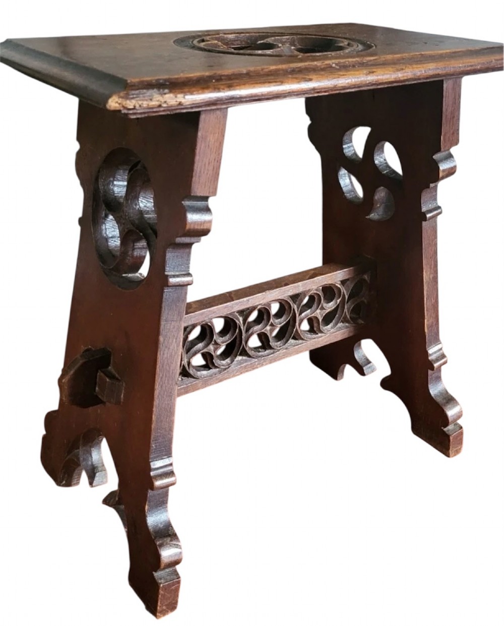 A 19th-century dark oak Gothic Revival stool featuring carved tracery cut-outs on the slab legs, a carved central stretcher, and a carved recessed motif on the top. View II