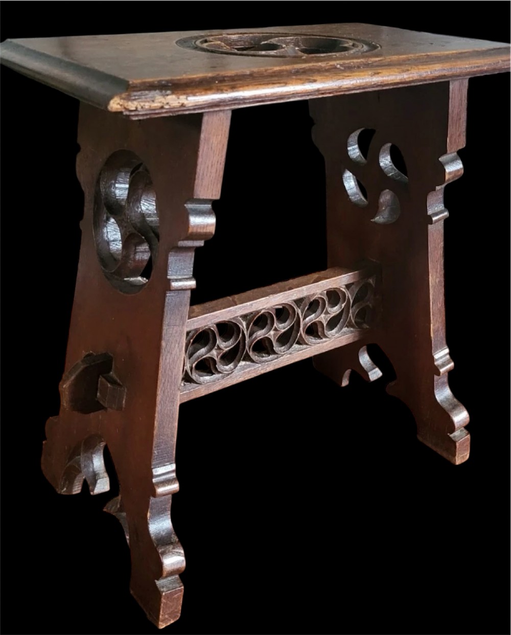 A 19th-century dark oak Gothic Revival stool featuring carved tracery cut-outs on the slab legs, a carved central stretcher, and a carved recessed motif on the top. View IV