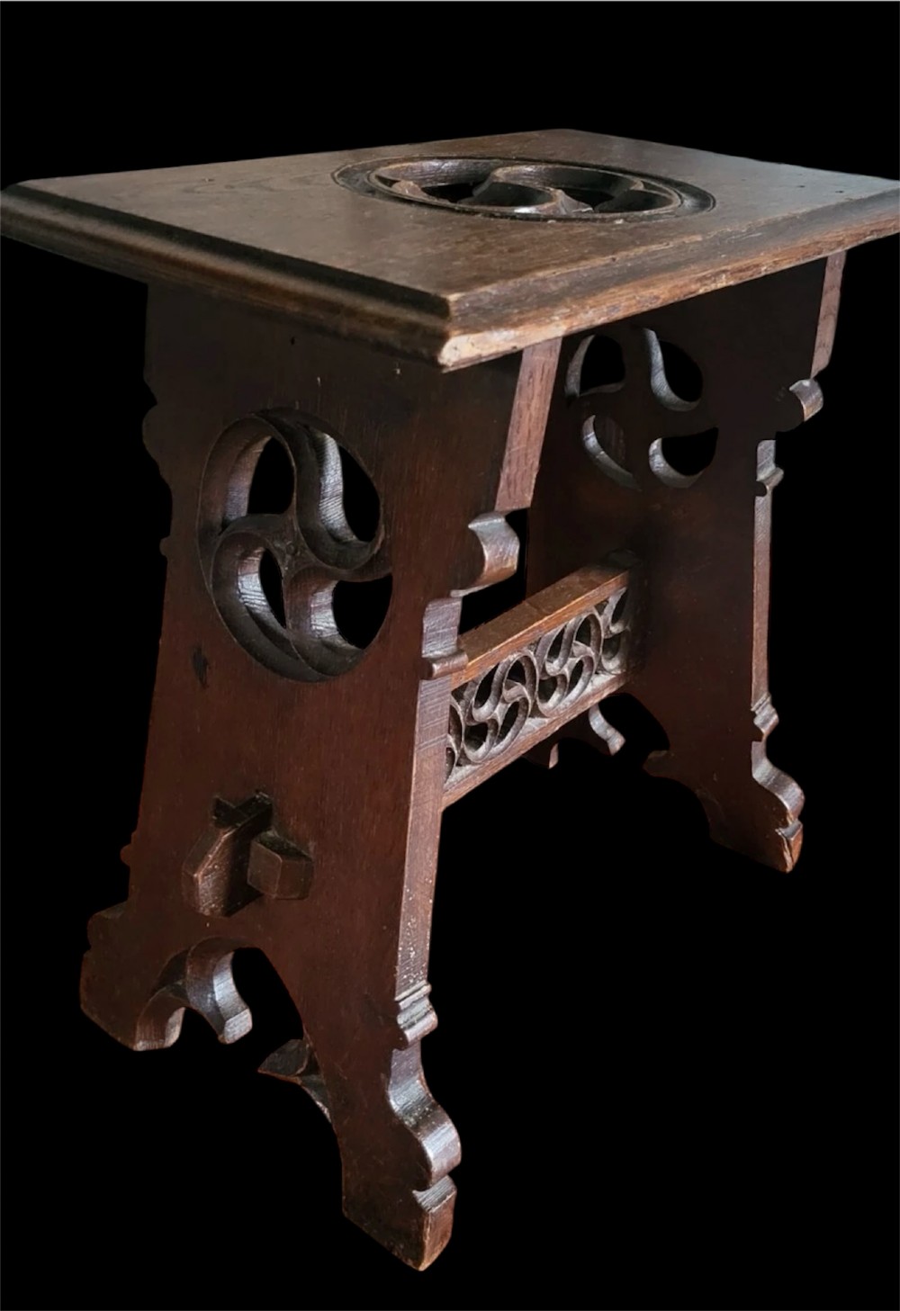 A 19th-century dark oak Gothic Revival stool featuring carved tracery cut-outs on the slab legs, a carved central stretcher, and a carved recessed motif on the top. View V