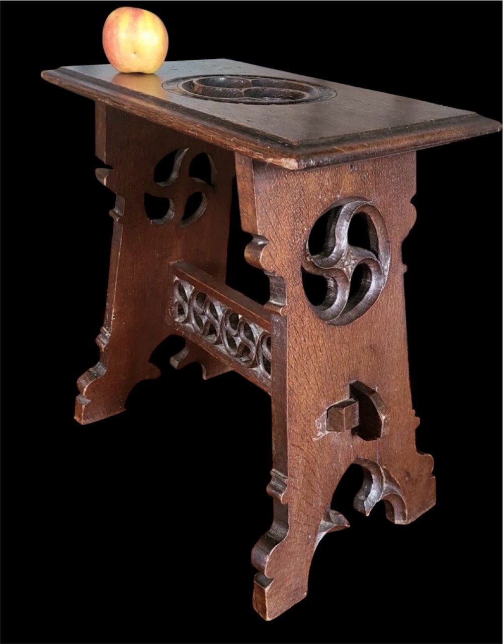 A 19th-century dark oak Gothic Revival stool featuring carved tracery cut-outs on the slab legs, a carved central stretcher, and a carved recessed motif on the top. View VI