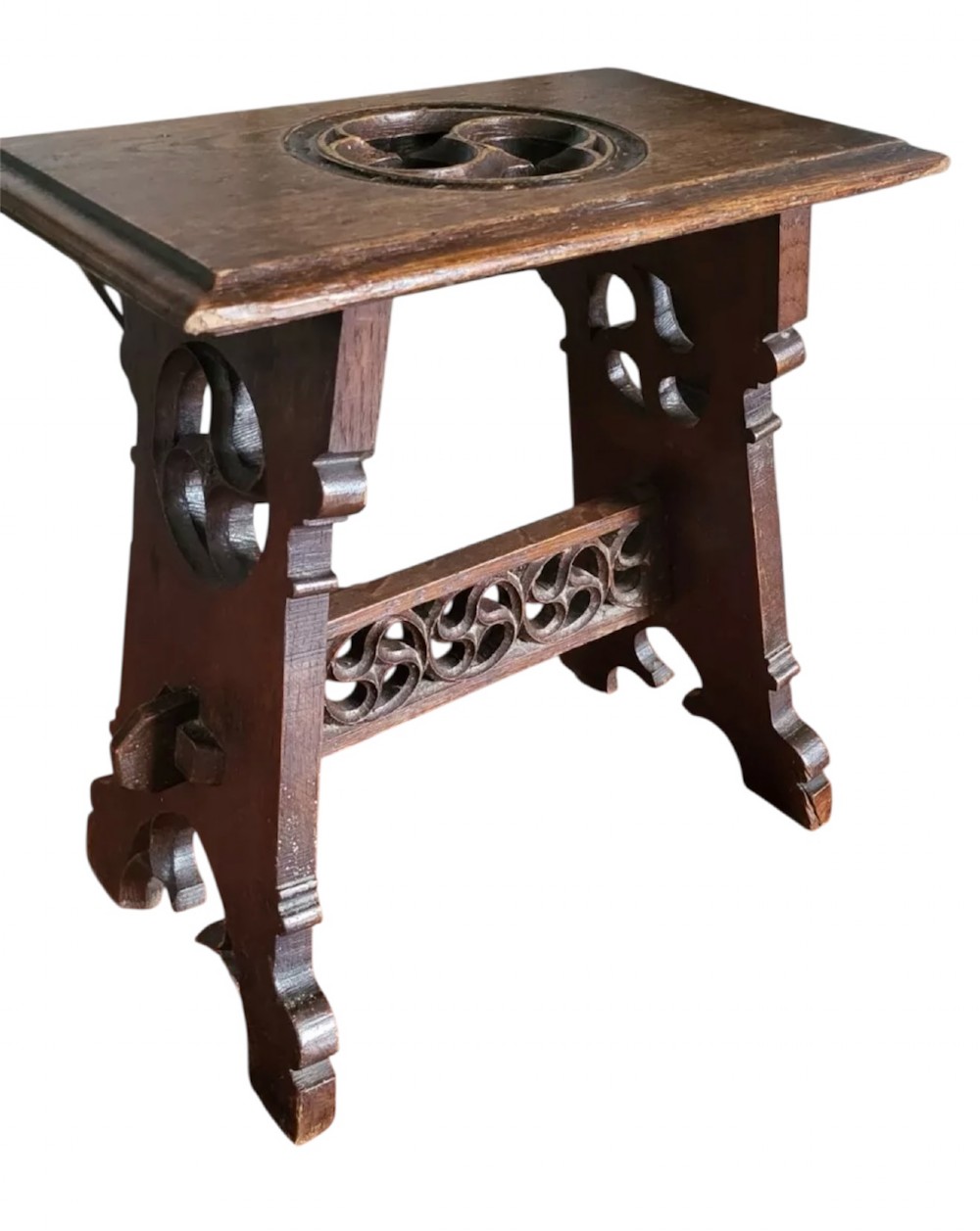 A 19th-century dark oak Gothic Revival stool featuring carved tracery cut-outs on the slab legs, a carved central stretcher, and a carved recessed motif on the top. View XIV