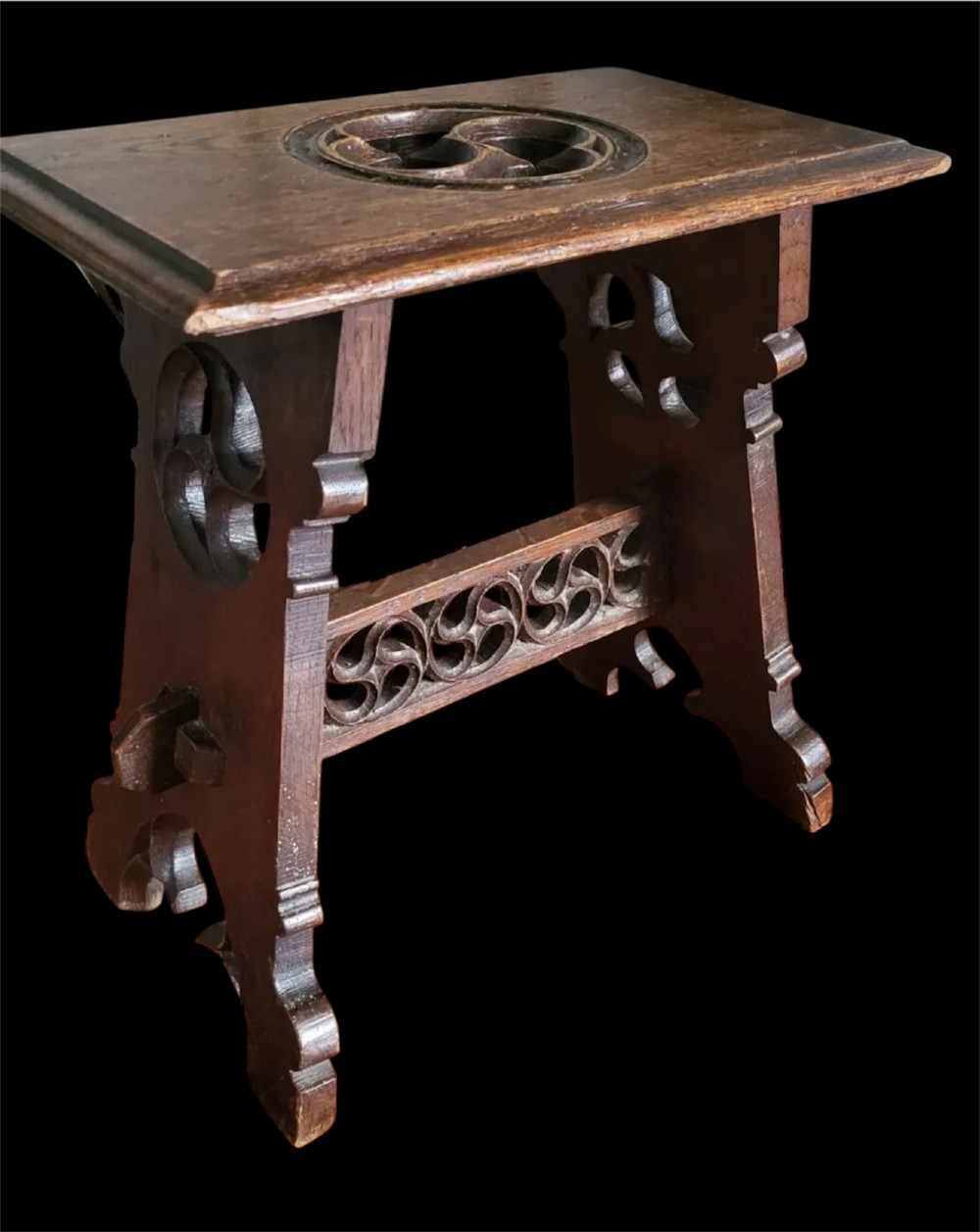 A 19th-century dark oak Gothic Revival stool featuring carved tracery cut-outs on the slab legs, a carved central stretcher, and a carved recessed motif on the top. View XV