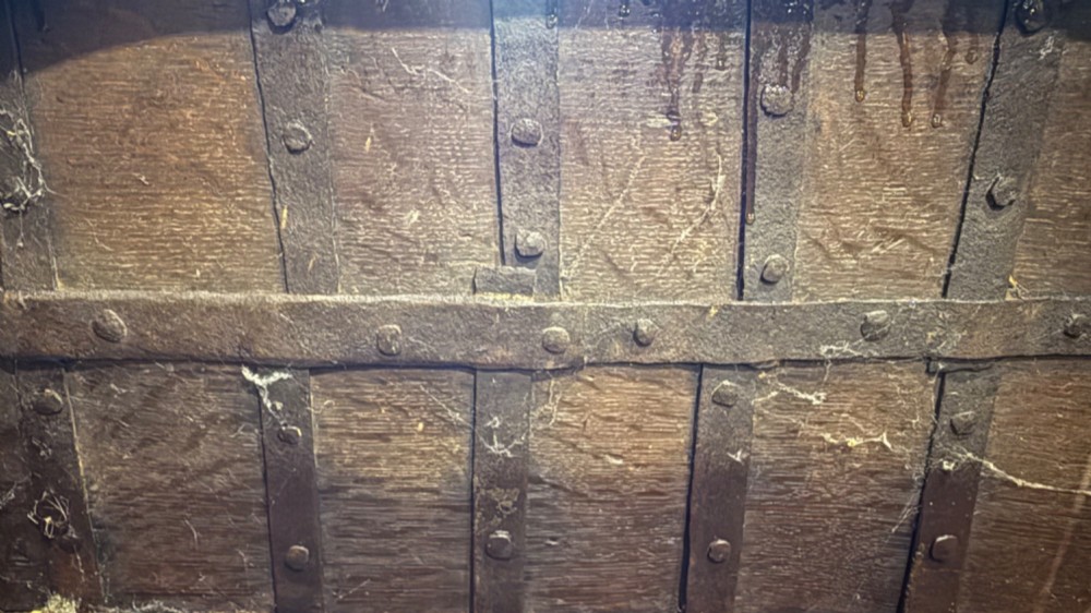 A sixteenth-century rectangular dark wood casket bound with riveted iron straps, featuring a top carrying handle and arched timber base. View XV