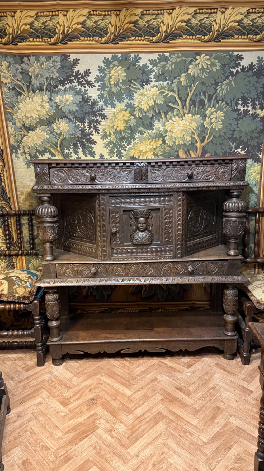 A 17th-century style dark carved oak livery cupboard featuring ornate relief carving, a central face motif panel, turned supports, and an open lower shelf.