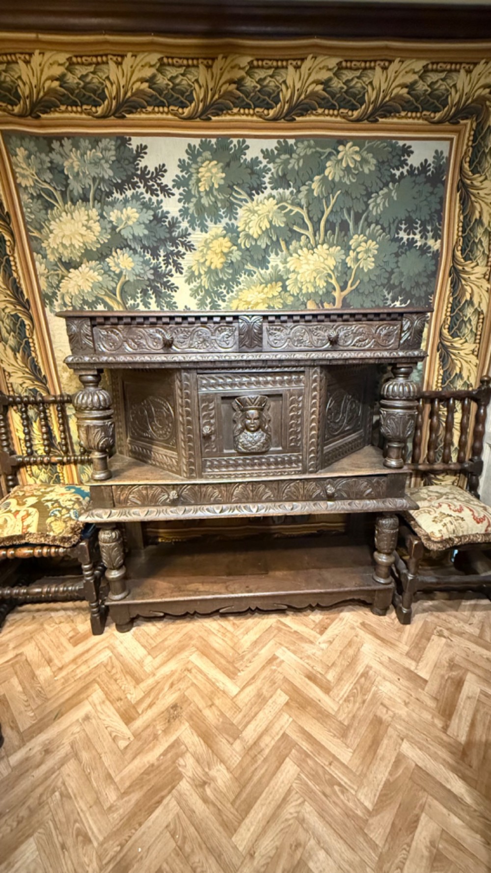 A 17th-century style dark carved oak livery cupboard featuring ornate relief carving, a central face motif panel, turned supports, and an open lower shelf. View II