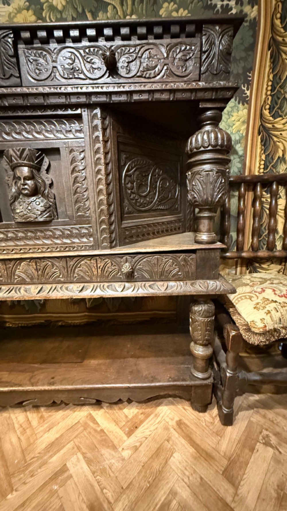 A 17th-century style dark carved oak livery cupboard featuring ornate relief carving, a central face motif panel, turned supports, and an open lower shelf. View III