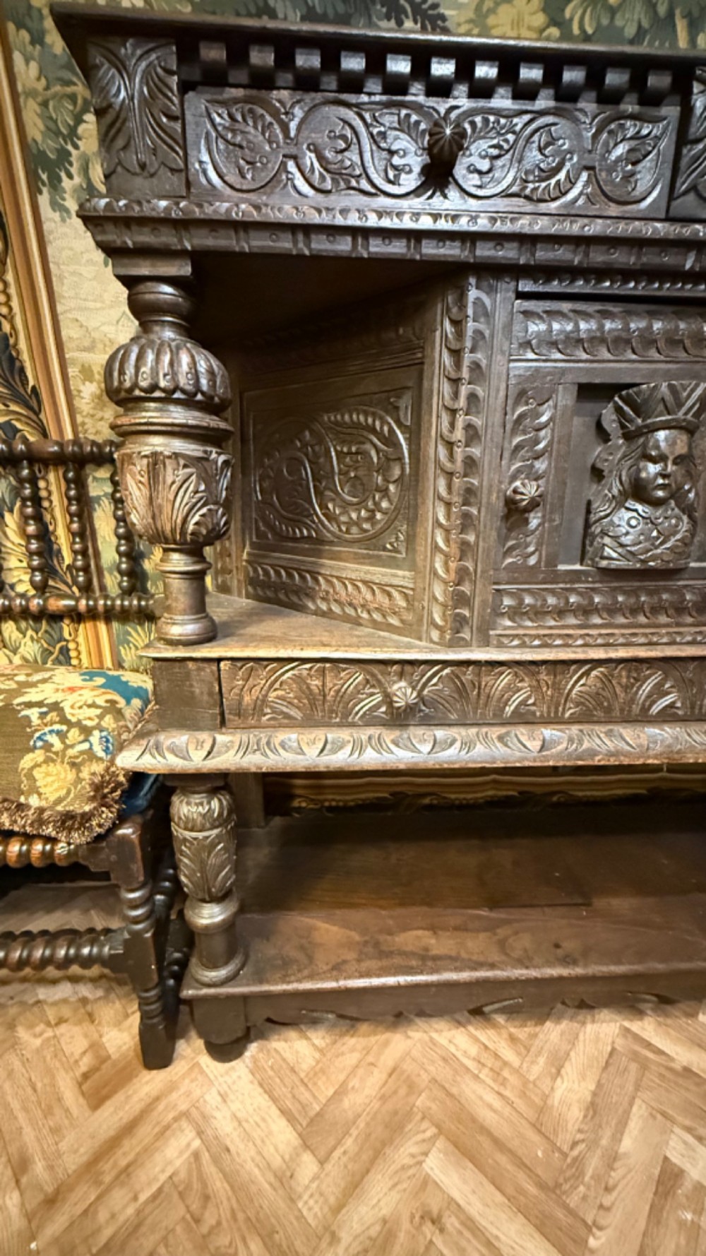 A 17th-century style dark carved oak livery cupboard featuring ornate relief carving, a central face motif panel, turned supports, and an open lower shelf. View IV