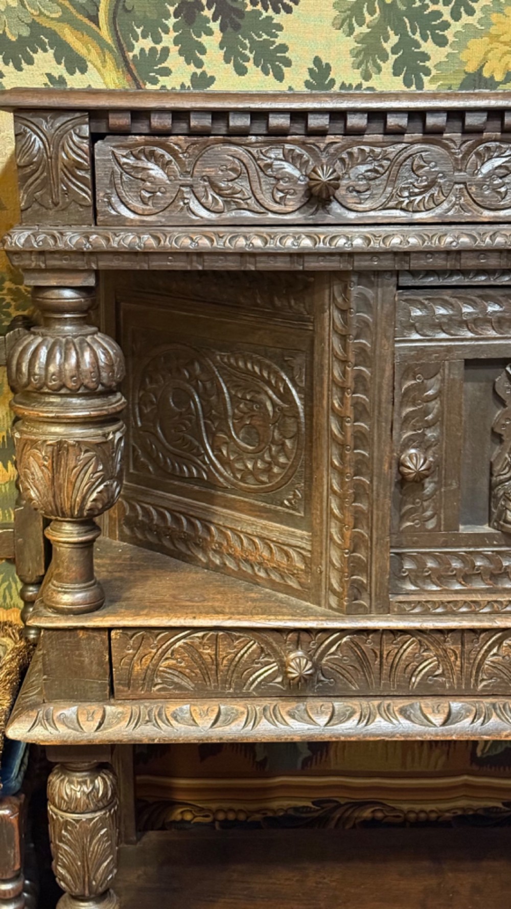 A 17th-century style dark carved oak livery cupboard featuring ornate relief carving, a central face motif panel, turned supports, and an open lower shelf. View VI