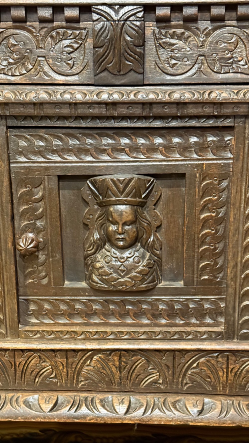 A 17th-century style dark carved oak livery cupboard featuring ornate relief carving, a central face motif panel, turned supports, and an open lower shelf. View IX