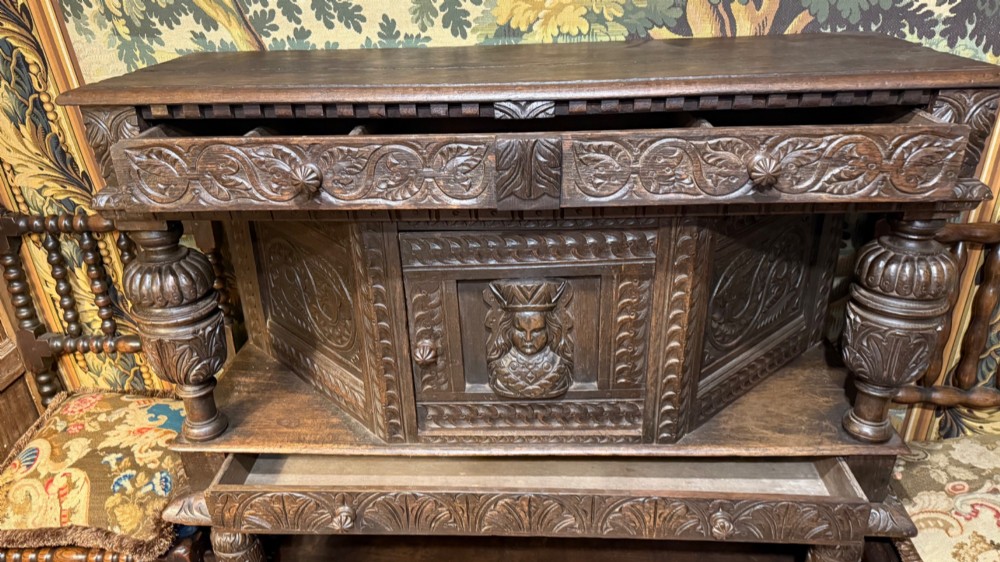 A 17th-century style dark carved oak livery cupboard featuring ornate relief carving, a central face motif panel, turned supports, and an open lower shelf. View XII