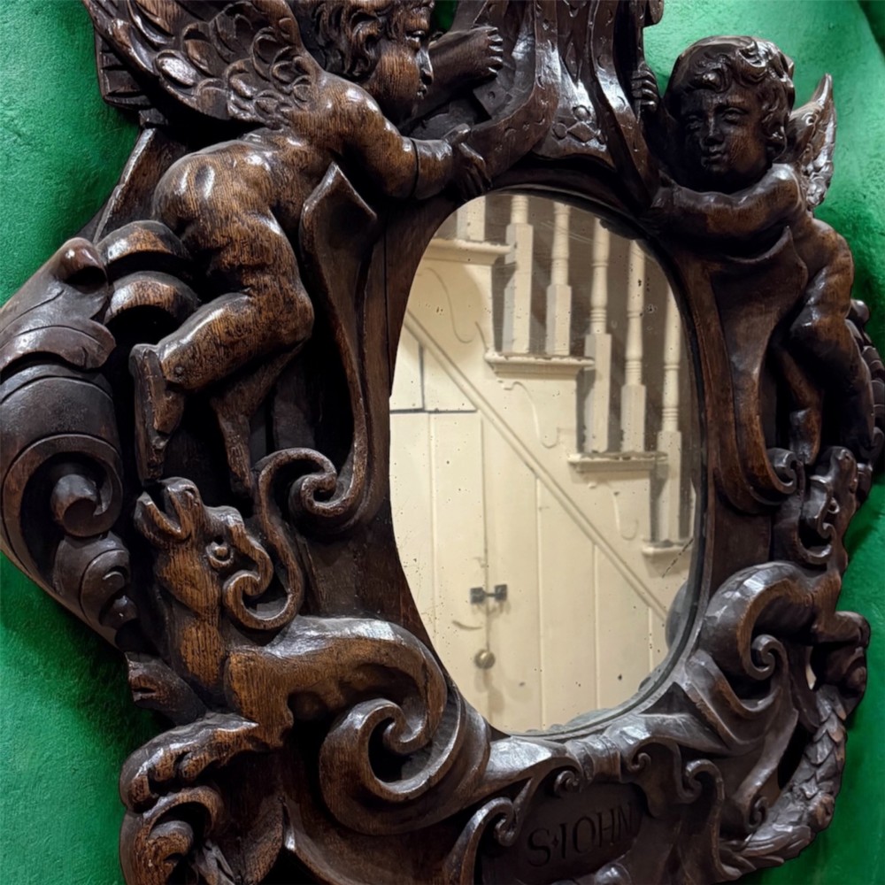 An 18th century dark carved oak wall mirror featuring carved cherubs and scrolling motifs framing an oval mirror plate. View II