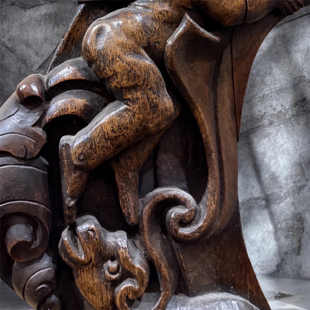 An 18th century dark carved oak wall mirror featuring carved cherubs and scrolling motifs framing an oval mirror plate. View IV