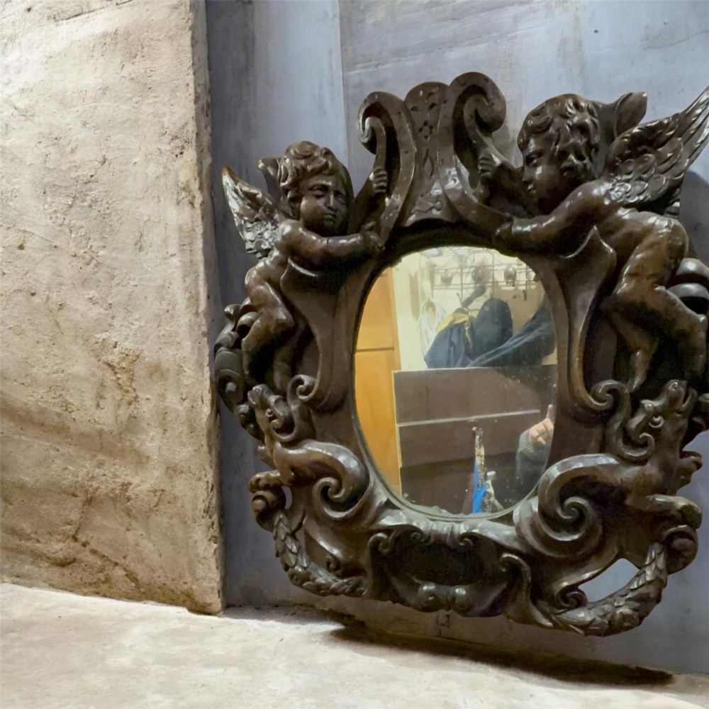 An 18th century dark carved oak wall mirror featuring carved cherubs and scrolling motifs framing an oval mirror plate.