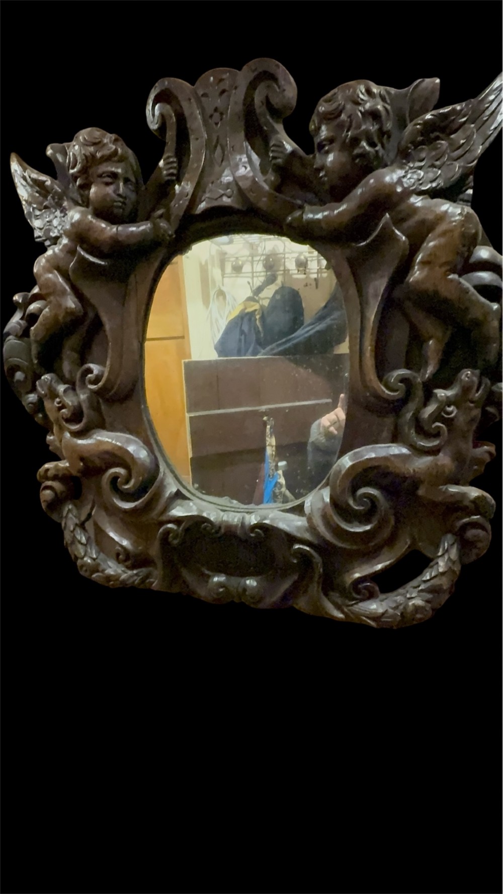 An 18th century dark carved oak wall mirror featuring carved cherubs and scrolling motifs framing an oval mirror plate. View VIII