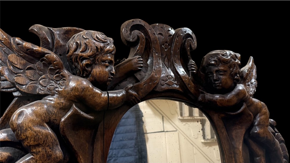 An 18th century dark carved oak wall mirror featuring carved cherubs and scrolling motifs framing an oval mirror plate. View X