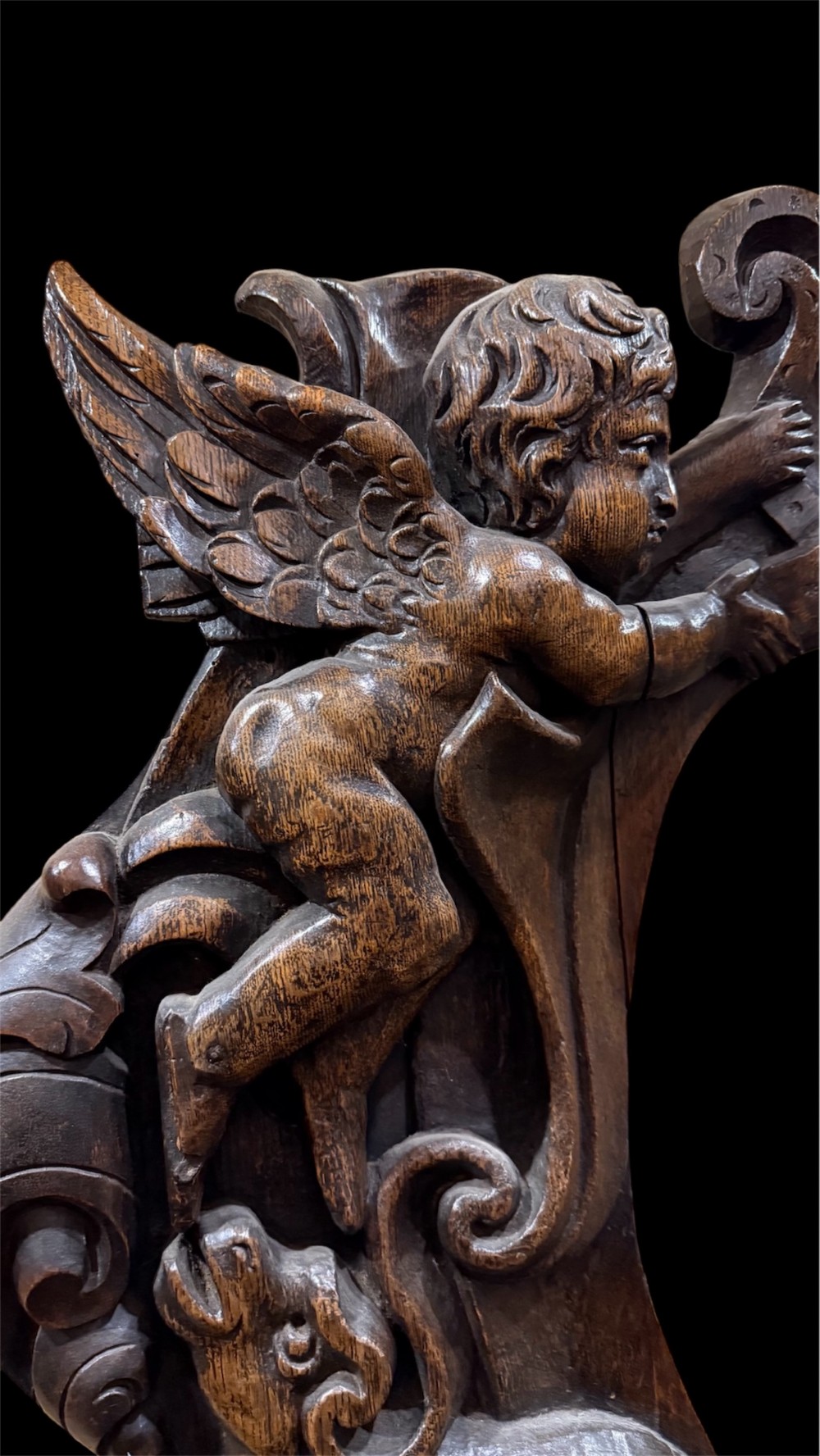 An 18th century dark carved oak wall mirror featuring carved cherubs and scrolling motifs framing an oval mirror plate. View XII