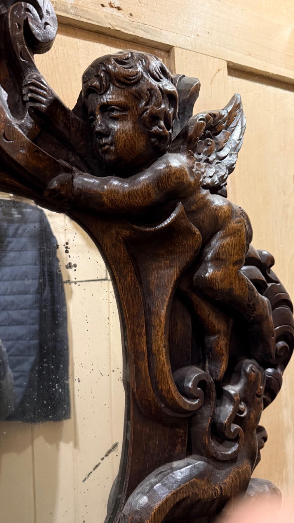 An 18th century dark carved oak wall mirror featuring carved cherubs and scrolling motifs framing an oval mirror plate. View XV