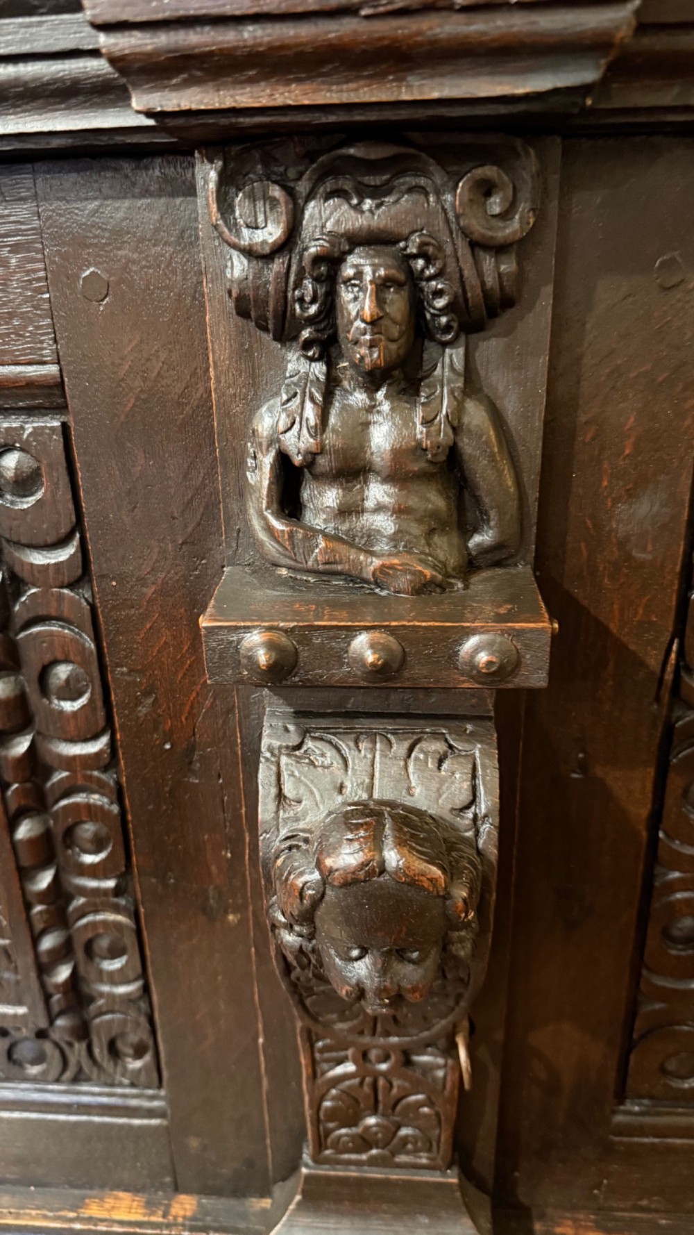 An 18th-century dark oak hanging cupboard featuring carved figural columns, relief door panels with floral motifs, and carved masks. View IX