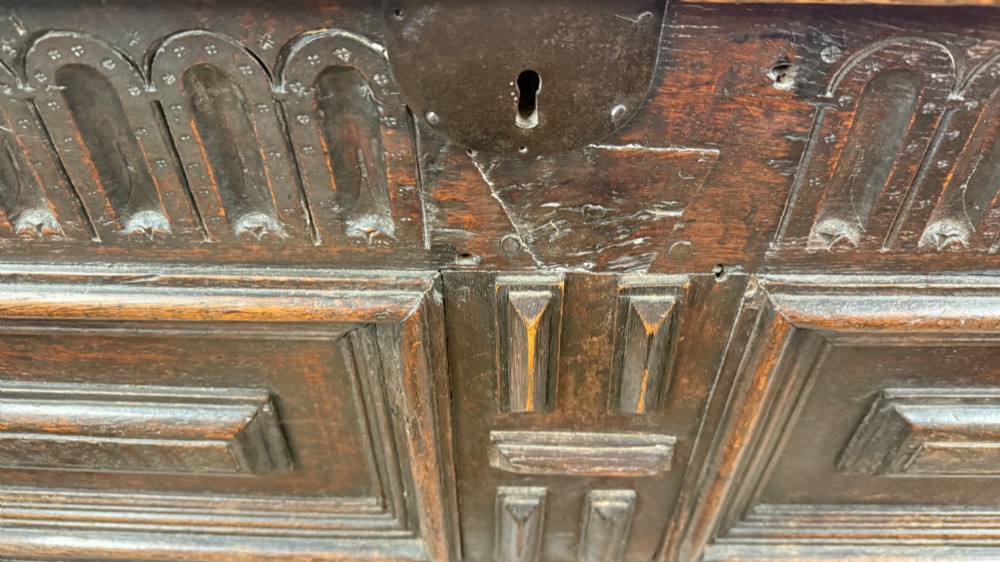 A 17th-century dark oak coffer featuring carved arcaded detail above geometrically moulded panel fronts and a central iron keyhole escutcheon. View VI