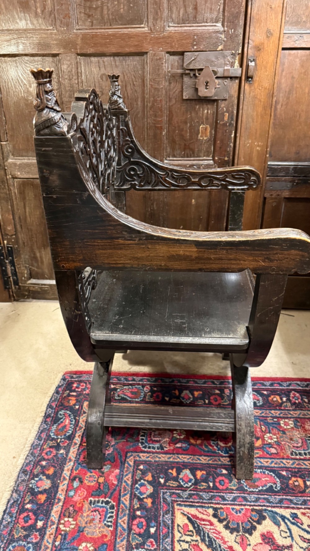 A 19th-century dark carved oak X-frame armchair featuring a foliate backrest, turned balusters, and carved head finials. View X