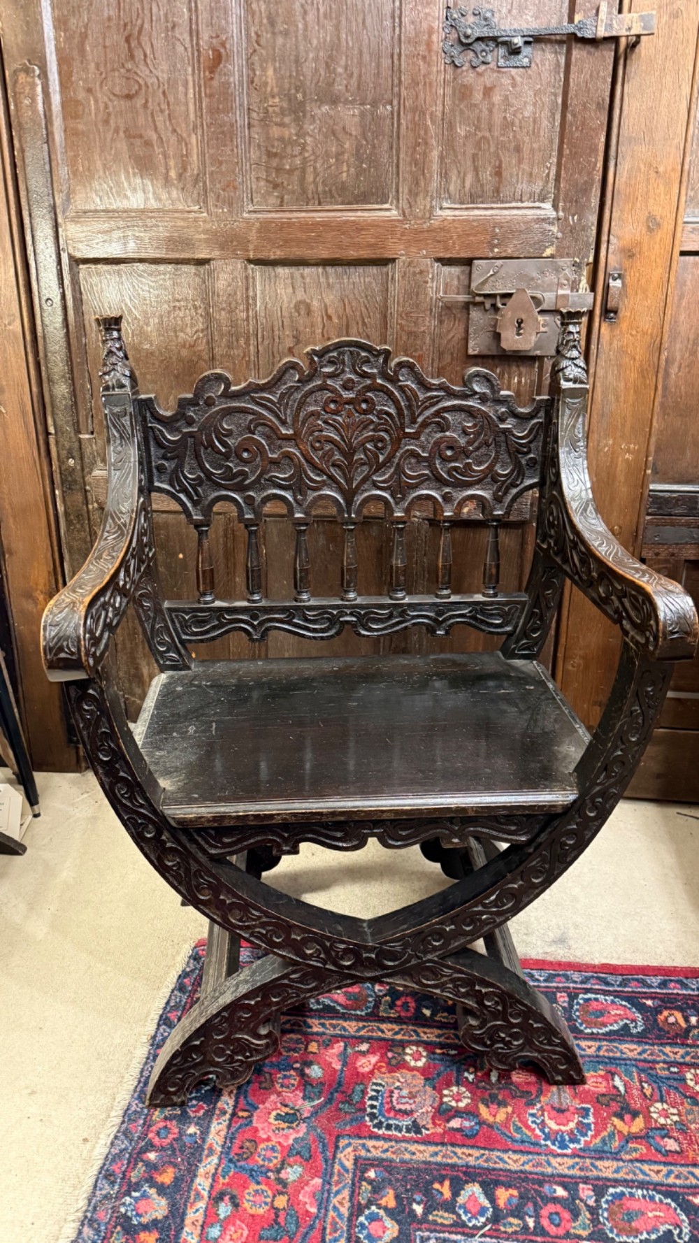 A 19th-century dark carved oak X-frame armchair featuring a foliate backrest, turned balusters, and carved head finials. View XI