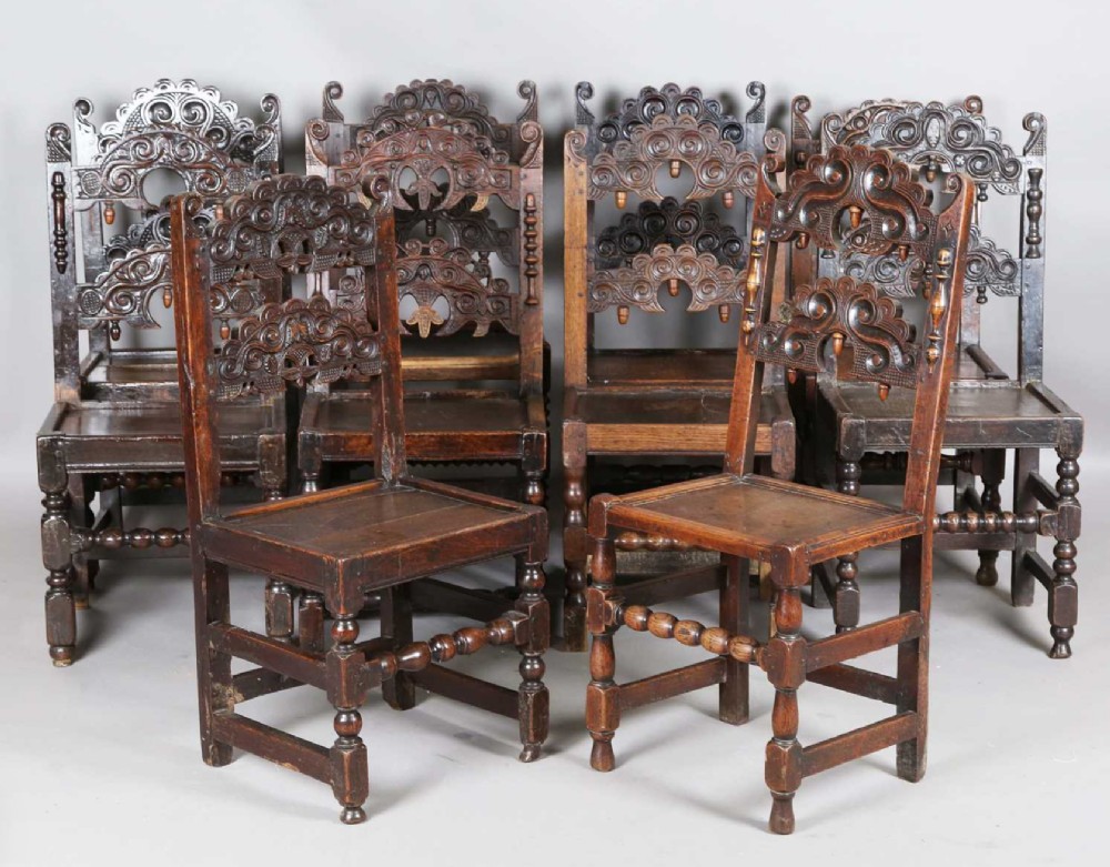 A set of dark carved oak dining chairs featuring carved openwork backs with scroll motifs, solid wooden seats, and turned front legs.