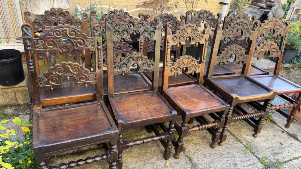 A set of dark carved oak dining chairs featuring carved openwork backs with scroll motifs, solid wooden seats, and turned front legs. View IX