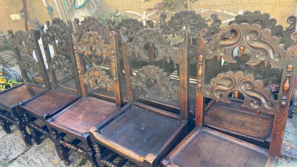 A set of dark carved oak dining chairs featuring carved openwork backs with scroll motifs, solid wooden seats, and turned front legs. View X