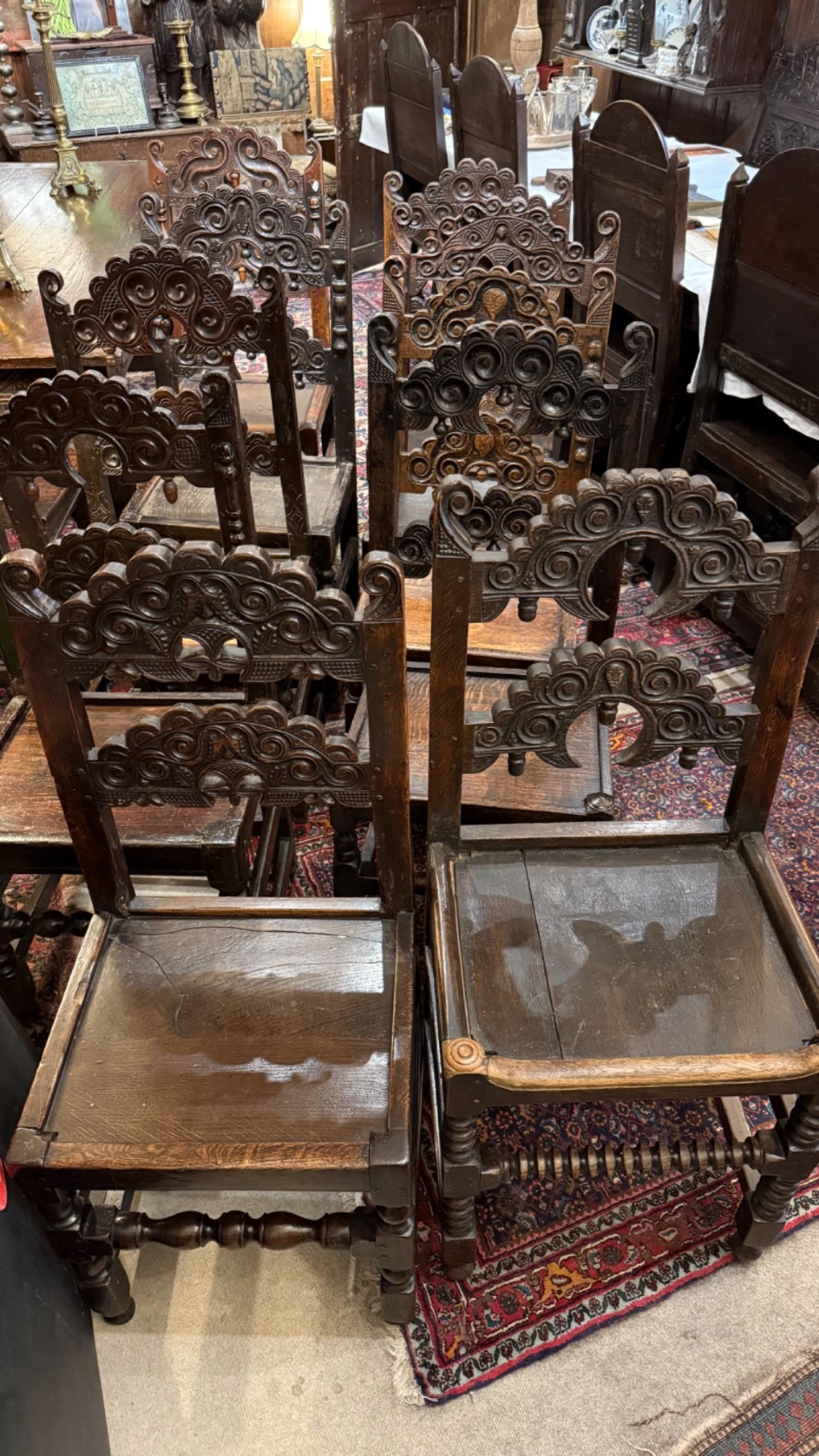 A set of dark carved oak dining chairs featuring carved openwork backs with scroll motifs, solid wooden seats, and turned front legs. View XI