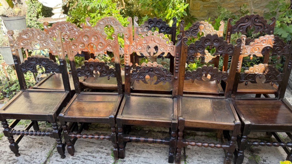 A set of dark carved oak dining chairs featuring carved openwork backs with scroll motifs, solid wooden seats, and turned front legs. View XII
