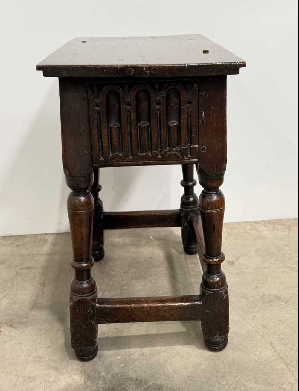 A 17th-century dark oak joint stool featuring a carved arcade frieze, turned legs, and sturdy base stretchers. View V