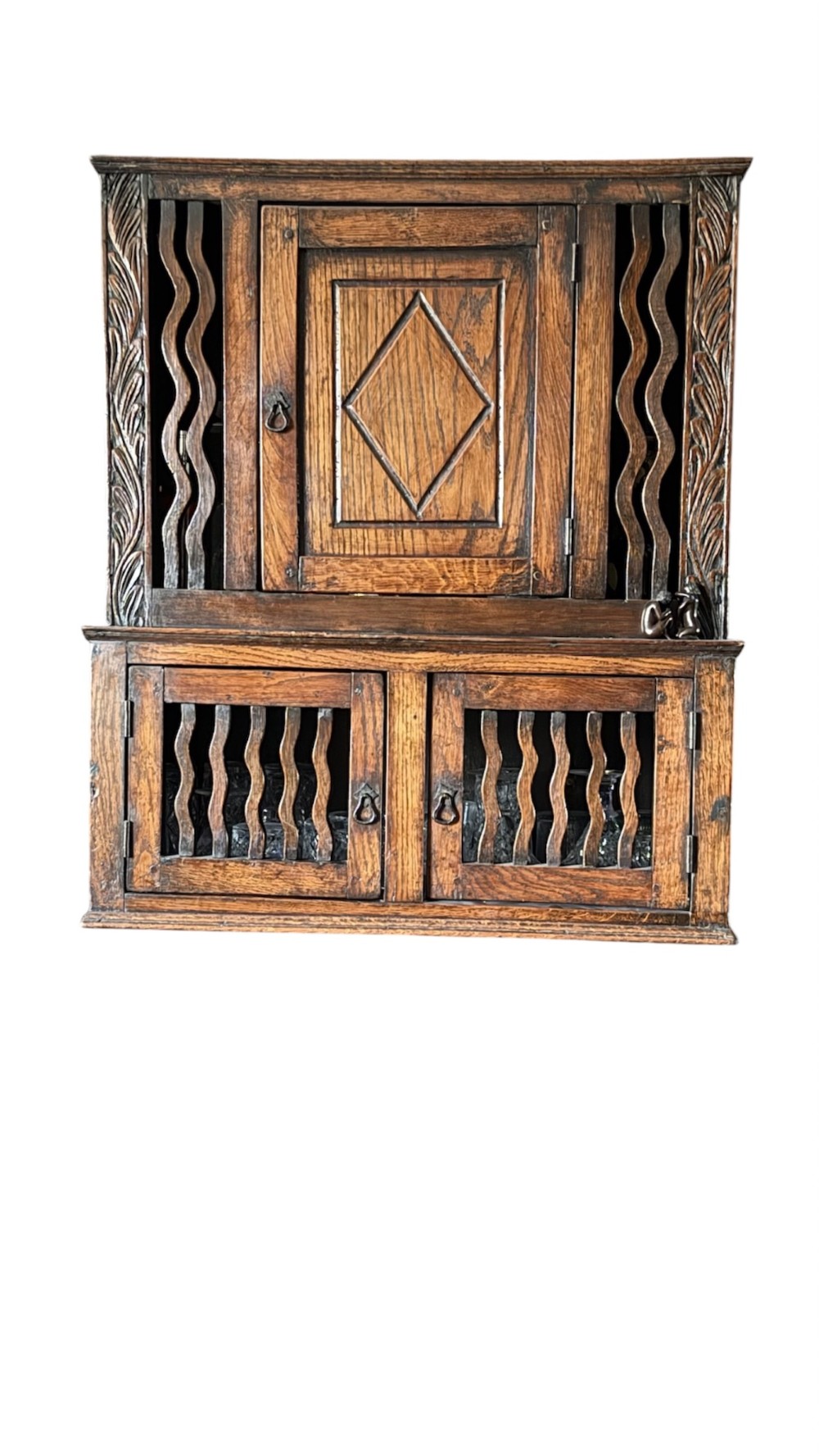 An 18th-century carved oak wall cabinet featuring a central door with a carved diamond motif flanked by wavy vertical slats and a lower two-door slatted cupboard. View II