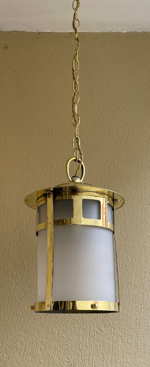 An Arts and Crafts style brass hall lantern with geometric cut-out metalwork and a frosted glass cylindrical shade, suspended by a brass chain. View II