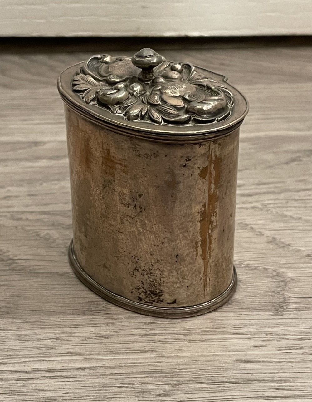 A cylindrical silver plated tea caddy featuring a repousse foliate patterned lid with a raised central finial. View II