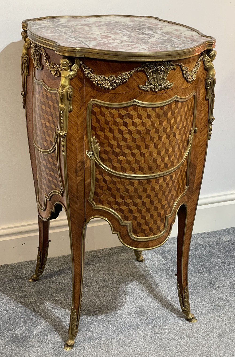 A French kingwood bedside cabinet featuring cube parquetry inlay, gilt ormolu mounts, floral garlands, and a shaped marble top.