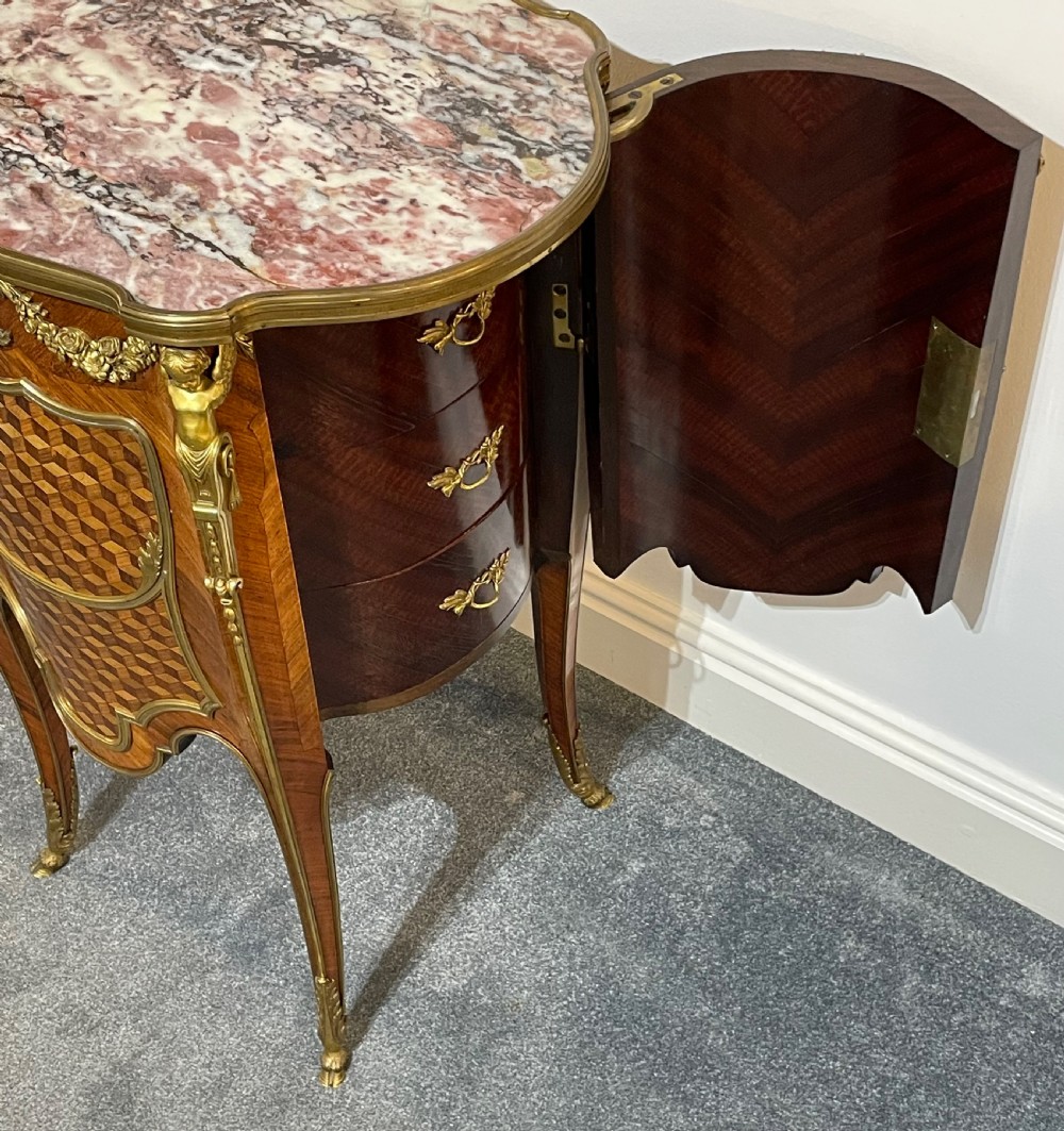 A French kingwood bedside cabinet featuring cube parquetry inlay, gilt ormolu mounts, floral garlands, and a shaped marble top. View X