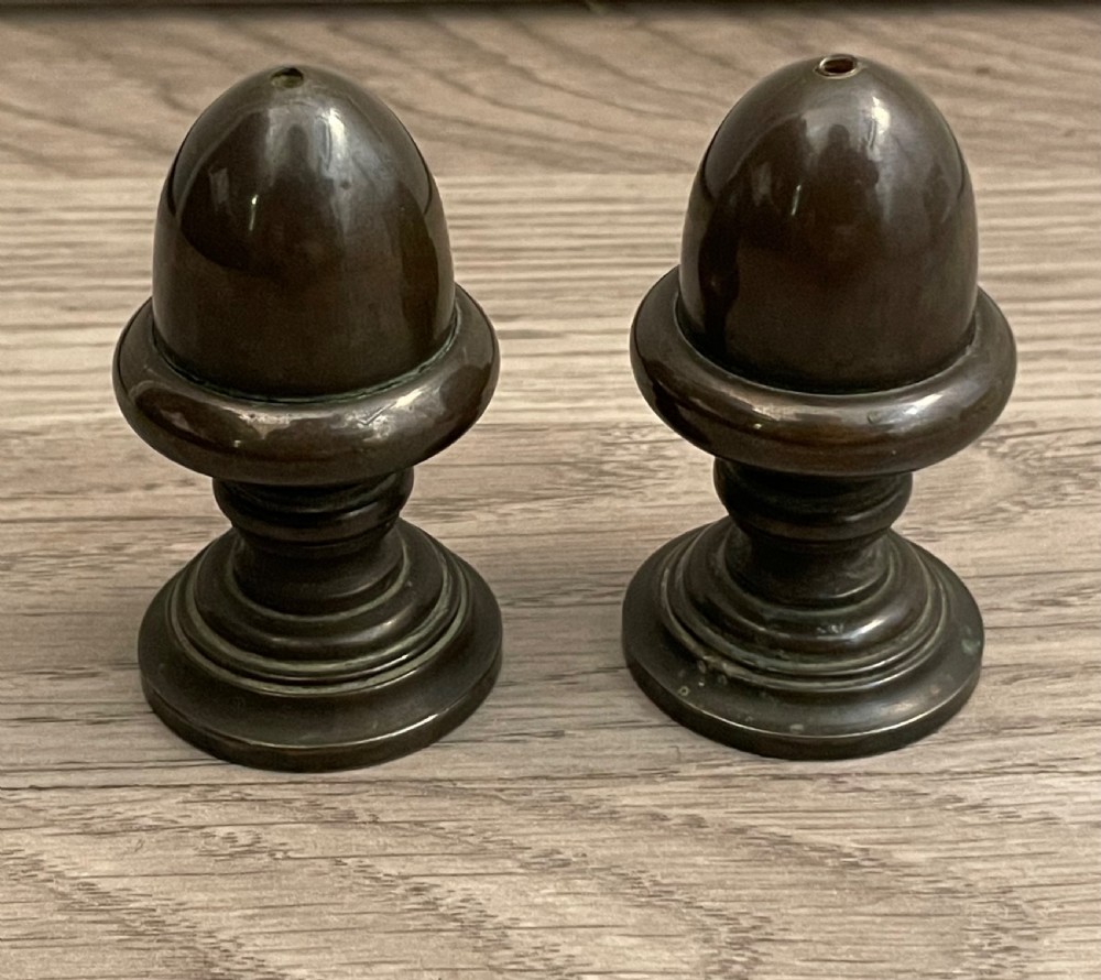 A pair of turned dark bronze spice shakers featuring acorn-form tops set upon circular stepped pedestal bases.