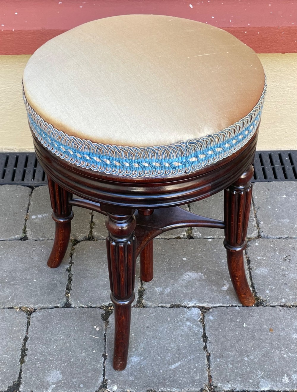 A circular Regency period revolving piano stool featuring a cushioned top with blue and cream braided trim, set upon four fluted, splayed legs in dark simulated rosewood.