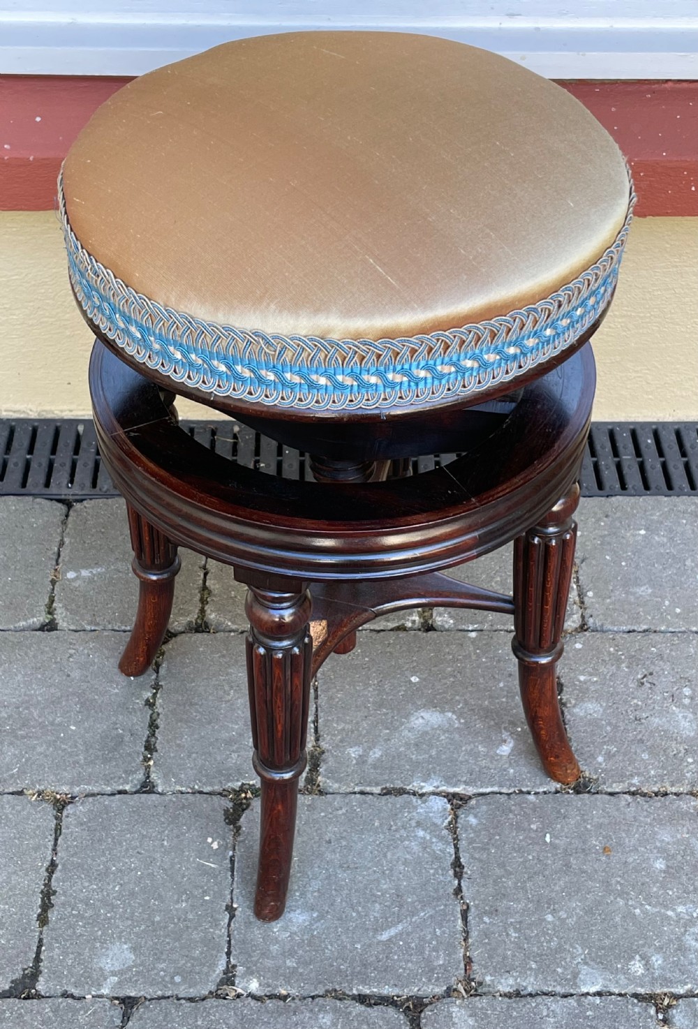 A circular Regency period revolving piano stool featuring a cushioned top with blue and cream braided trim, set upon four fluted, splayed legs in dark simulated rosewood. View II