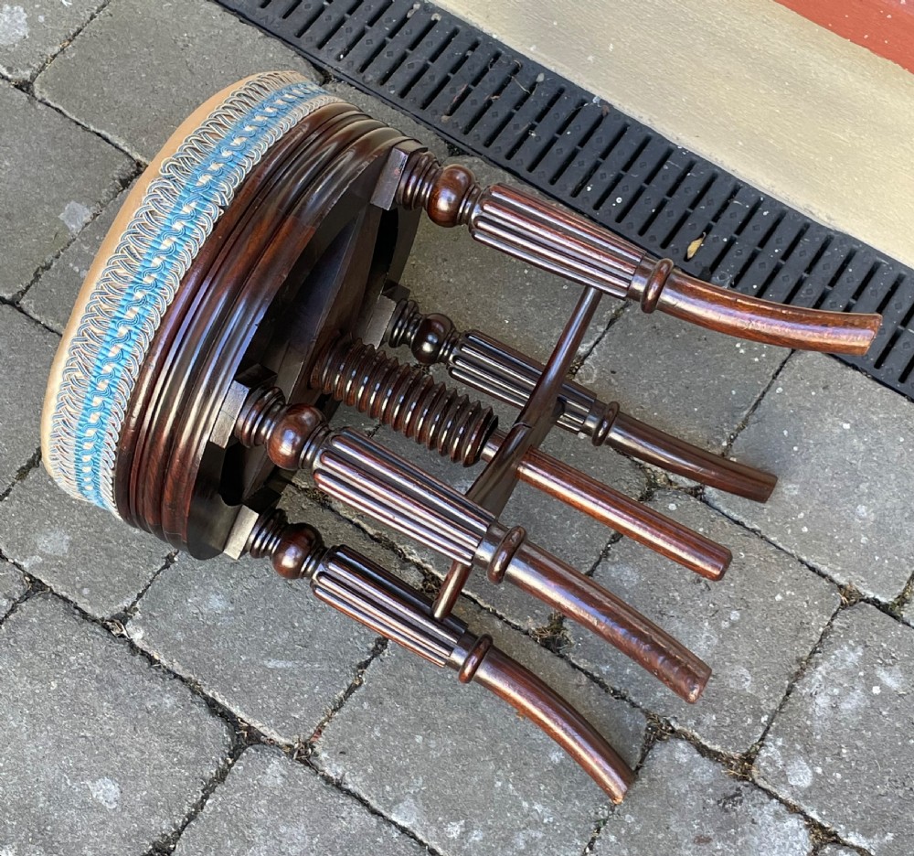 A circular Regency period revolving piano stool featuring a cushioned top with blue and cream braided trim, set upon four fluted, splayed legs in dark simulated rosewood. View V