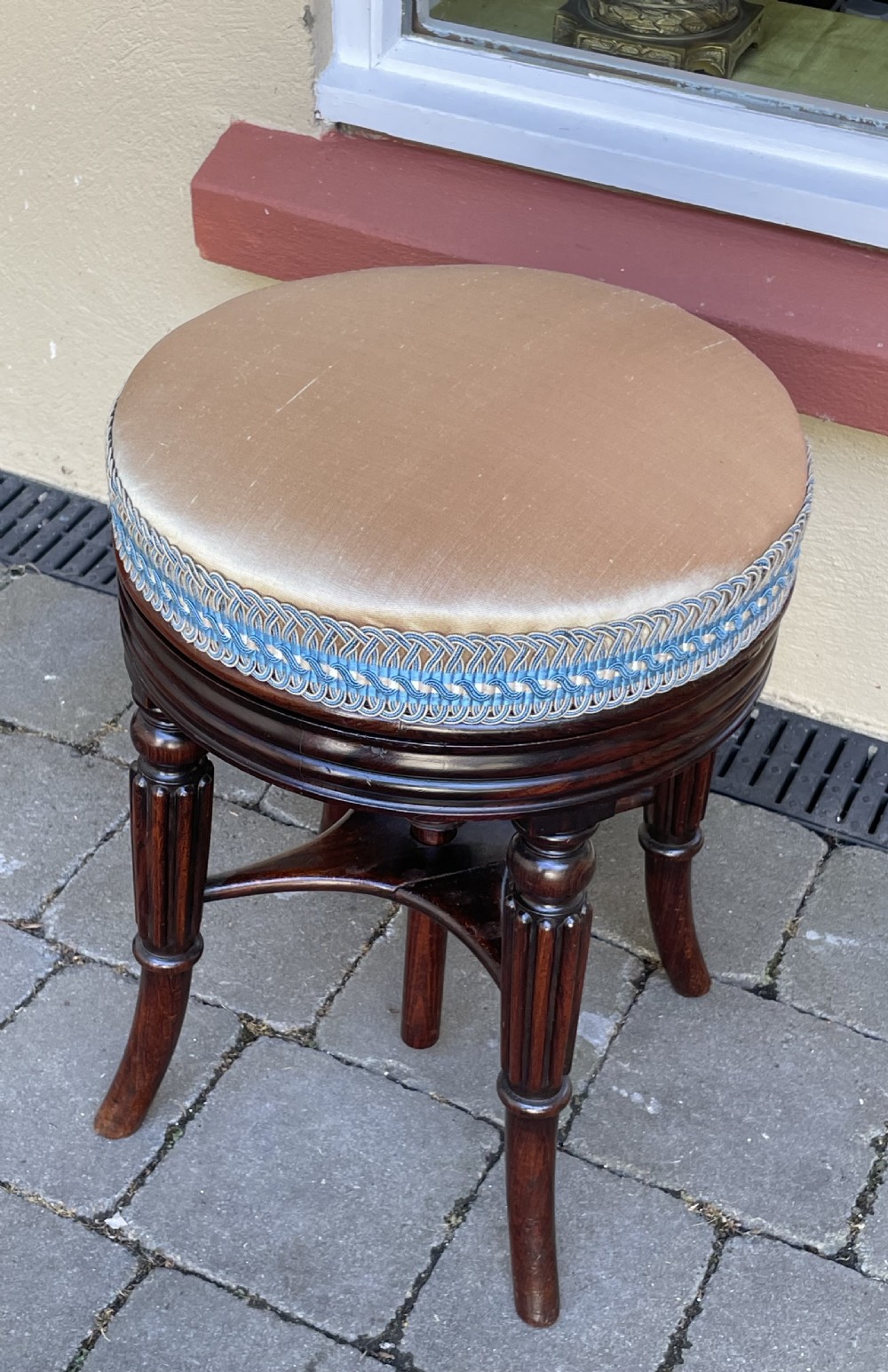 A circular Regency period revolving piano stool featuring a cushioned top with blue and cream braided trim, set upon four fluted, splayed legs in dark simulated rosewood. View VII